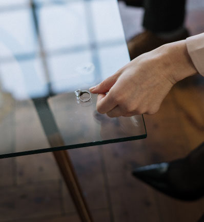 A person's hand reaching towards a solitary engagement ring on a glass surface, with reflections and wooden flooring in the background, capturing a moment of decision.