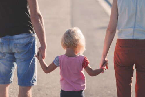 A small child holding hands with two adults, walking together as a family on a sunlit road.