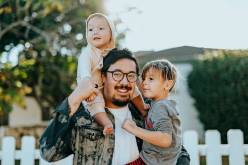 A joyful father with a toddler on his shoulders and a young child by his side, sharing a happy moment outdoors by a white picket fence, reminiscent of the diverse families represented by Nassau County LGBTQ