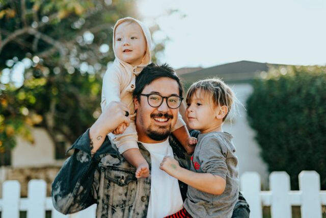 A joyful father with a toddler on his shoulders and a young child by his side, sharing a happy moment outdoors by a white picket fence, reminiscent of the diverse families represented by Long Island LGBTQ