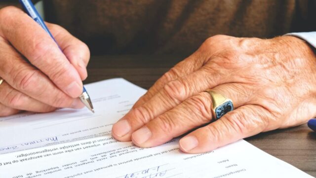 Close-up of a person's hands signing a Long Island Prenuptial Agreement, with attention to the details of aged skin and a ring on the right hand, emphasizing the act of agreement or