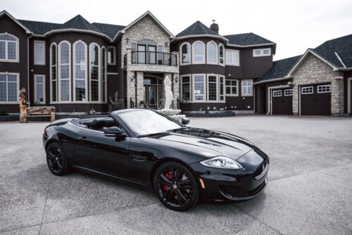 A sleek black convertible sports car, potentially discussed in a same sex divorce case in New York, parked in the driveway of a luxurious and expansive two-story residence with multiple garages and stone detailing.