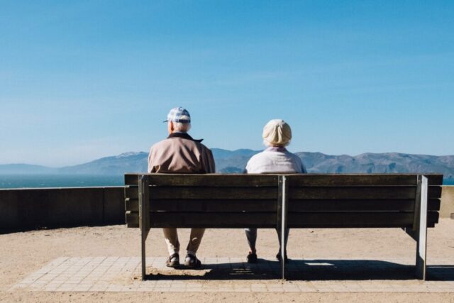 Two individuals sitting side by side on a park bench, gazing into the distance at a scenic mountainous landscape under a clear blue sky, pondering their next steps with a Jewish divorce lawyer.