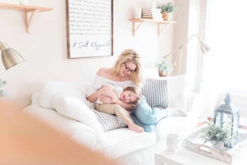 A warm and cozy moment with a smiling woman and child cuddling on a white sofa in a sunlit room decorated in a soft, neutral palette, reminiscent of the peace one might seek while consulting with
