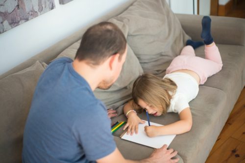 During a child visitation, a young child focused on drawing with colored pencils while lying on a couch, with an adult sitting beside her, observing her artwork.