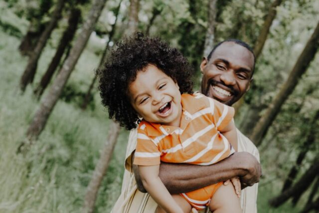 A joyful father holding his laughing toddler, sharing a playful and happy moment outdoors surrounded by greenery, embodying the serenity before facing complex challenges like same-sex divorce in New York.