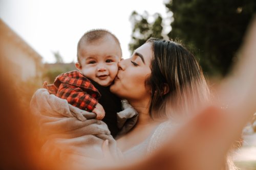 A mother giving her baby a tender kiss on the cheek, with the baby looking directly at the camera and smiling. They are captured in a warm, outdoor setting with soft sunlight filtering through. This serene