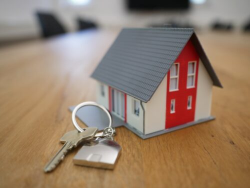 A set of keys with a keychain lies on a wooden surface in the foreground, symbolically suggesting home ownership or real estate, with a small model house in soft focus in the background, evoking