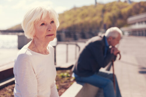 An elderly woman sitting outdoors with a contemplative expression, reflecting on her gray divorce, while a man rests with his walking stick in the background on a sunny day.