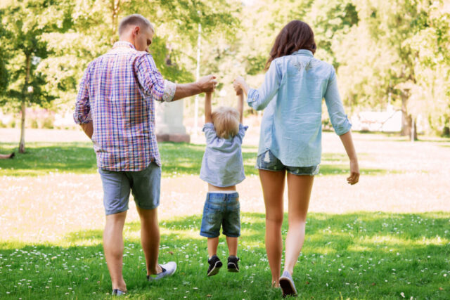A family enjoys a sunny day in the park, with a toddler from a remarrying couple taking steps while holding hands with his parents.