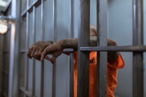 A person in an orange prison uniform stands behind metal jail bars, their tattooed arms resting through the bars—a stark portrayal of incarceration, with their face mostly obscured.