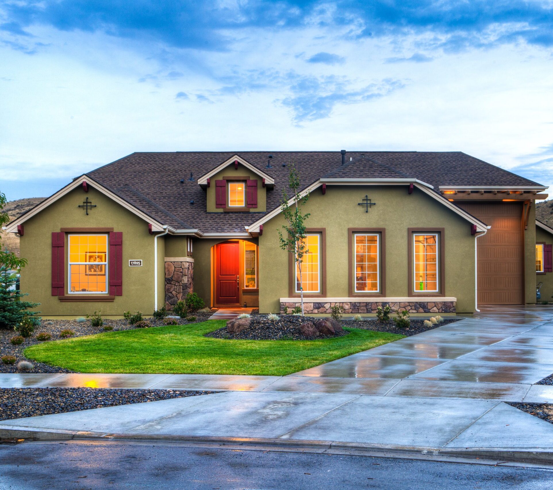 A single-story house with yellow-green walls, red trim, and large windows glows warmly at dusk. A Nassau County Real Estate Attorney can help guide you through buying a home like this, complete with a wet driveway, green lawn, and landscaped shrubs and trees.