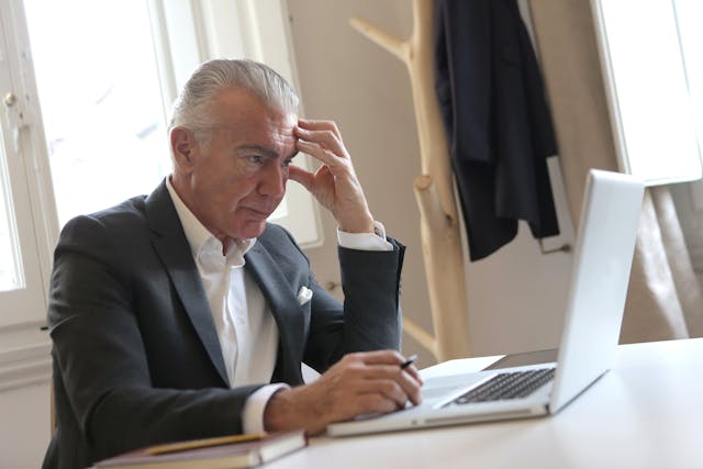An older man in a suit sits at a desk, looking thoughtfully at a laptop screen with his hand on his forehead. A notebook and pen are in front of him, and a coat hangs on a stand in the background.