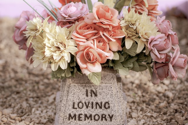 Artificial flowers in soft pink and cream shades are arranged in a vase attached to a small gray memorial stone engraved with "IN LOVING MEMORY." The background is covered with pebbles, creating a peaceful space for funeral instructions or quiet reflection.