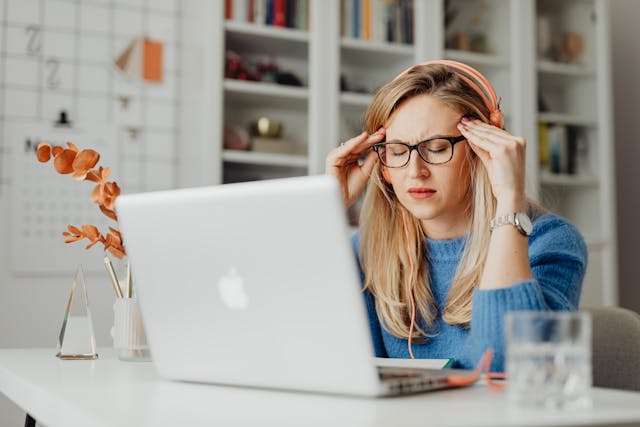 A woman wearing headphones and glasses sits at a desk with a laptop, looking stressed as she navigates her pro se divorce. She holds her temples, surrounded by a glass of water, notebook, and decorative items; shelves line the background.