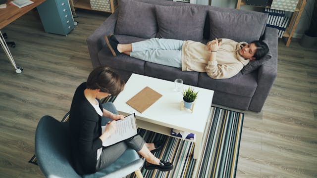 A child lies on a gray couch during a child's therapy session, while a therapist sits nearby, taking notes on a clipboard. A coffee table with a plant and papers sits between them.