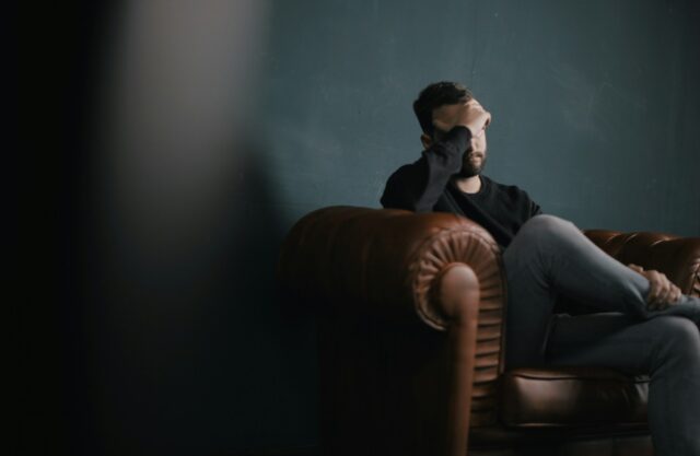 A man in New York sits on a brown leather sofa, legs crossed, resting his elbow on the armrest and covering his face with his hand, appearing stressed as he thinks about rehabilitating from addiction to regain custody.