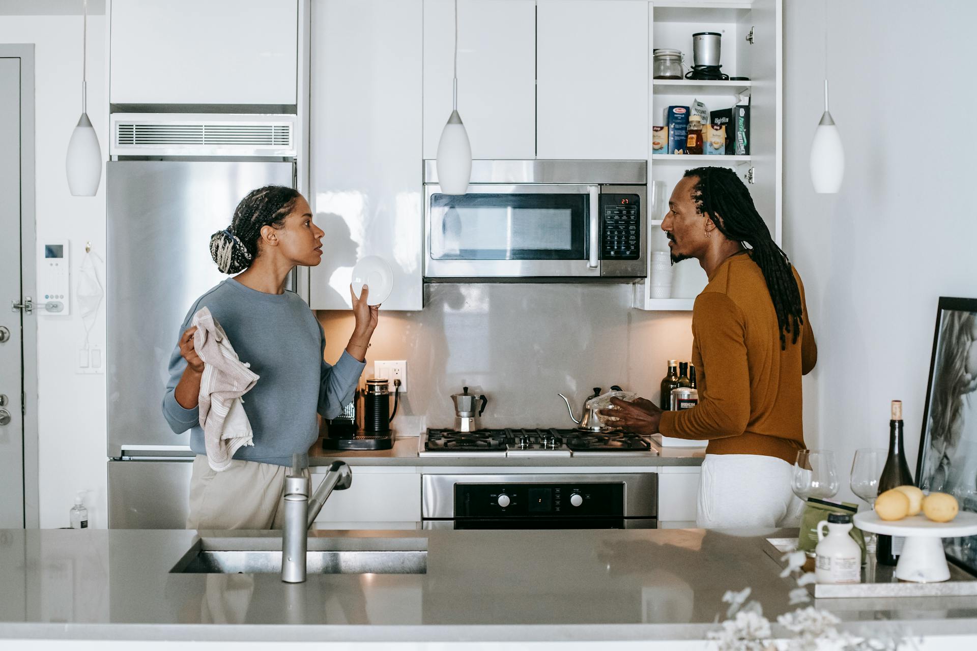 Two people stand in a modern kitchen having a serious conversation about the waiting period for divorce. One holds a dish and towel, while the other gestures with their hands. Kitchen appliances and shelves with items are visible in the background.