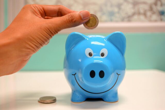 A hand holds a coin above a blue piggy bank with a smiling face, ready to drop it in—an act of careful saving akin to the diligence of a forensic accountant. Another coin rests on the white table; the background is gently blurred.