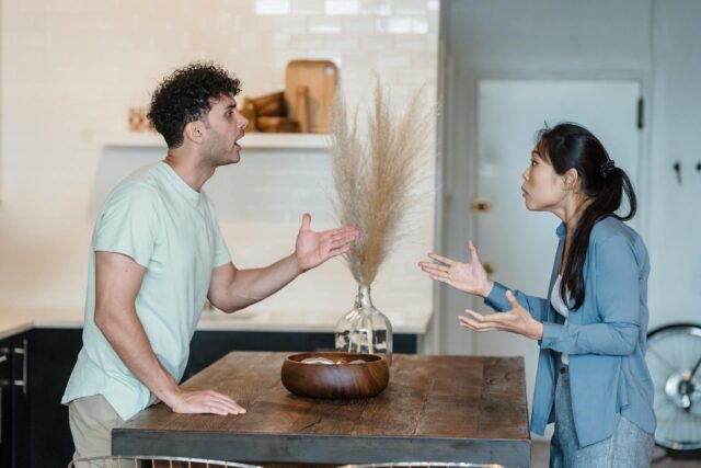 A man and woman are standing on opposite sides of a wooden kitchen table, facing each other and having an intense argument, gesturing with their hands—as if caught in a high conflict divorce. The modern kitchen has white tiles and stylish decor.