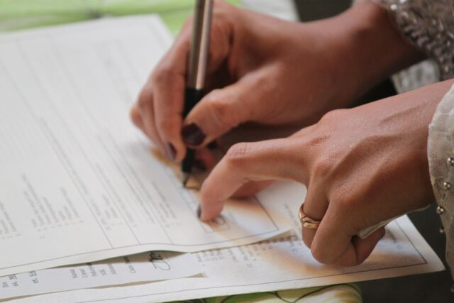 A close-up of a person’s hands holding a pen and signing a prenuptial agreement, with another sheet of paper beneath—possibly related to child custody. The person is wearing a ring and a garment with decorative beads.