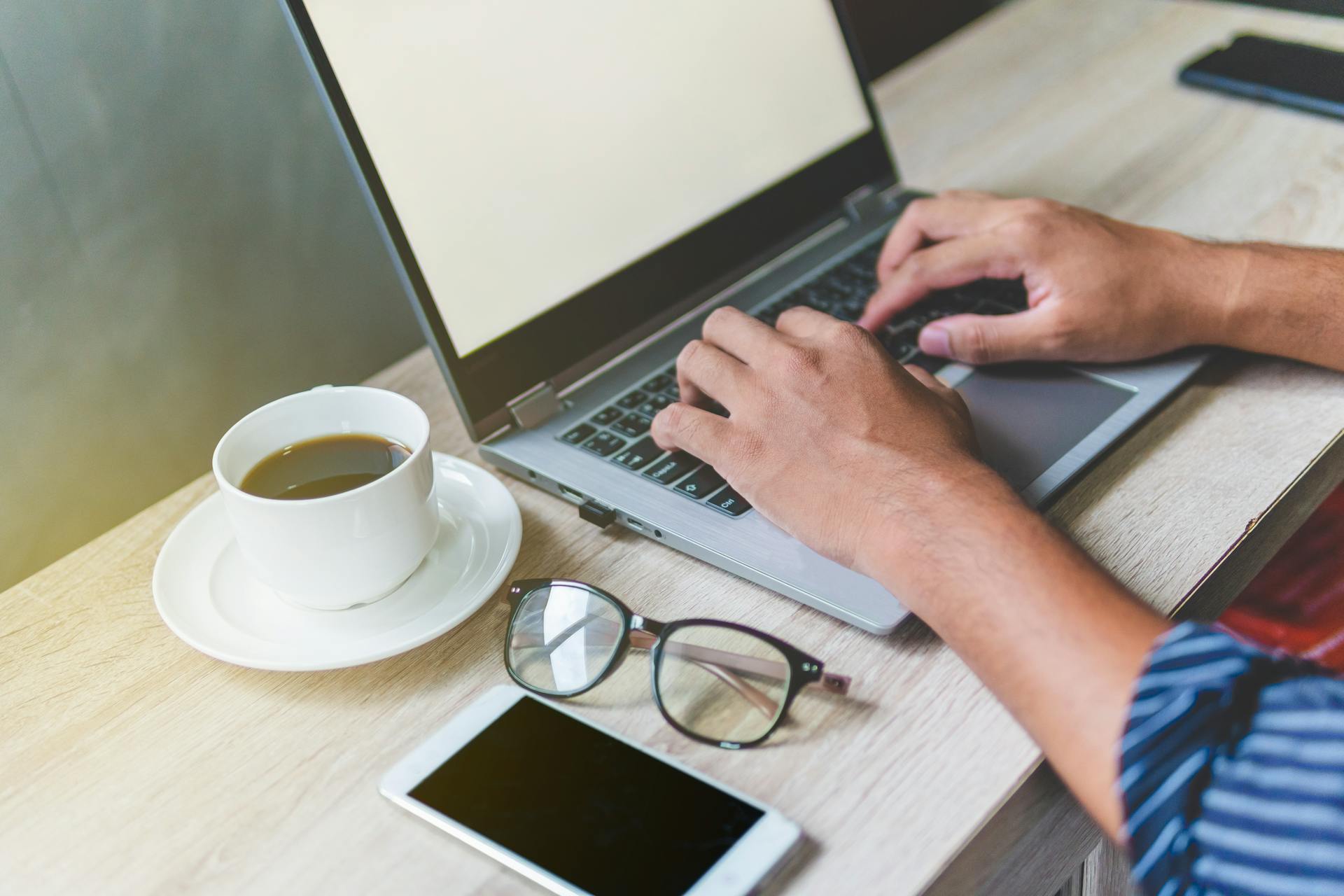 A person typing on a laptop at a wooden desk, creating a will, with a cup of coffee, eyeglasses, and a smartphone placed nearby.