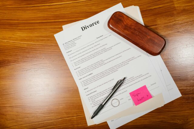 A divorce document sits on a wooden table with a black pen, a brown eyeglass case, and a pink sticky note reading “sign here” attached—awaiting signatures despite the spouse missing from the scene.
