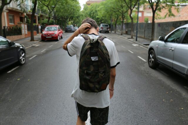 A teenager with a camouflage backpack walks down an empty street, scratching their head. Cars are parked along both sides, and trees line the sidewalks on a cloudy day.