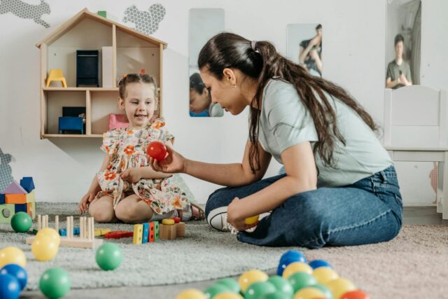 A woman and a young girl sit on the floor playing with colorful balls and toys in a playroom. The girl smiles while the woman, keeping the Best Interest of the Child in mind, hands her a red ball. A dollhouse and mirrors are visible in the background.