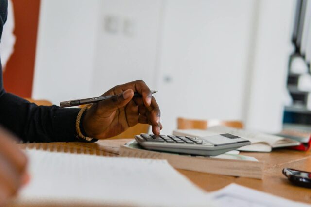 A person holding a pen and pressing buttons on a calculator sits at a desk with an open notebook, focusing on taxes or studying in a bright room.