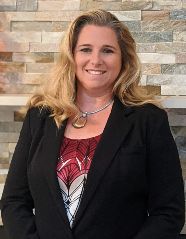 Diana Szabo, Esq. A professional woman, who is a Long Island child support lawyer, smiling for a portrait, wearing a black blazer and a red patterned blouse, with a stone pendant necklace, in front of