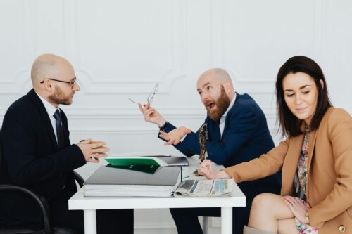 Three people in business attire sit around a white table with documents, a newspaper, and a small statuette. Two men are engaged in a lively discussion over a divorce petition, while a woman sits nearby, looking down and smiling slightly.