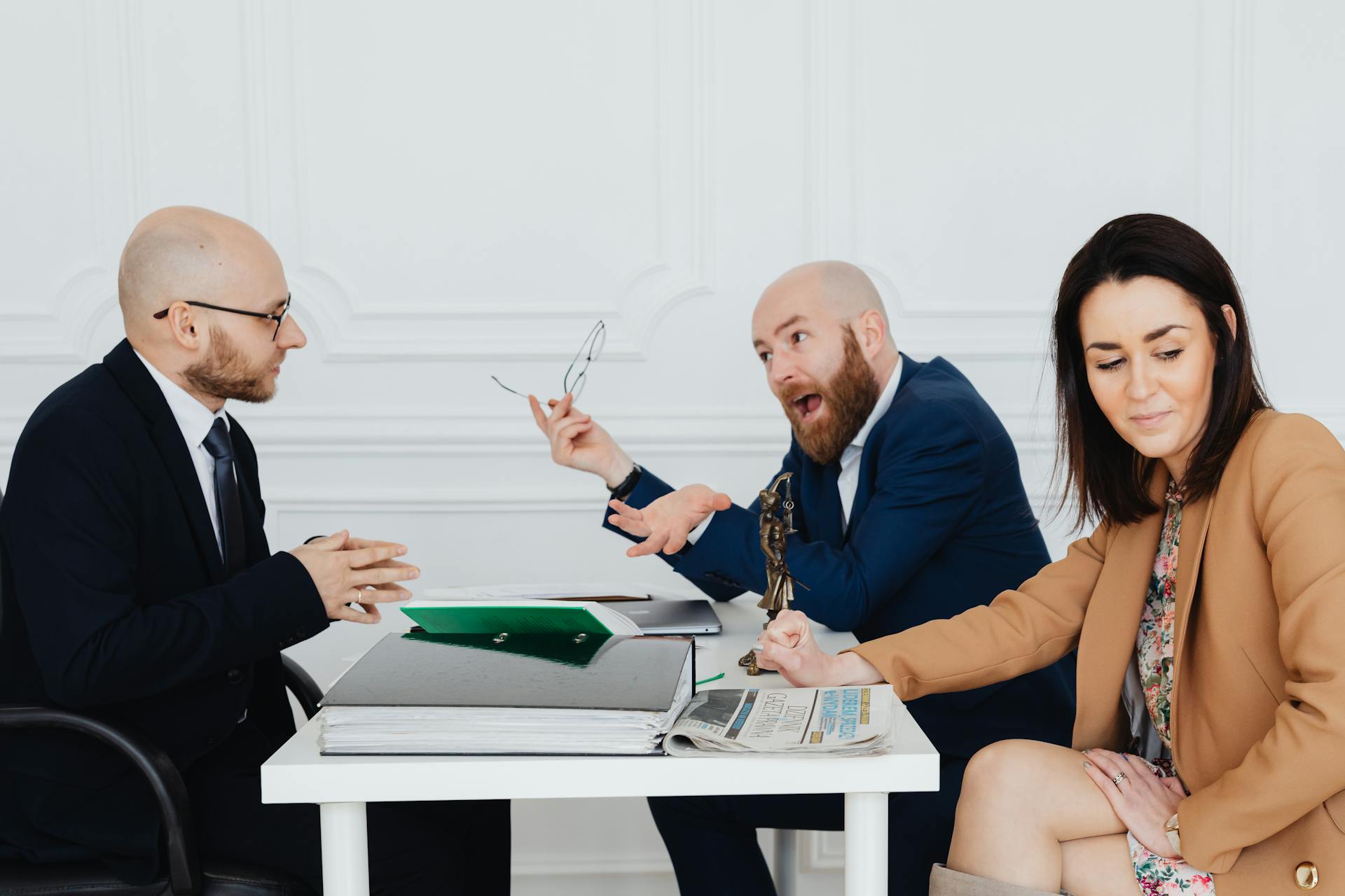 Three people in business attire sit around a white table with documents, a newspaper, and a small statuette. Two men are engaged in a lively discussion over a divorce petition, while a woman sits nearby, looking down and smiling slightly.